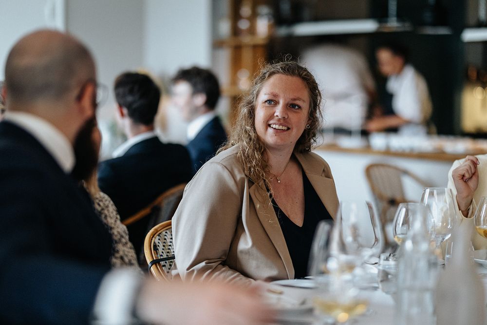 A guest smiles warmly while dining at an upscale restaurant with wine glasses and formal table settings.