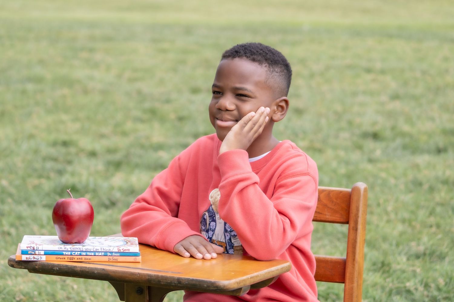 Student in coral sweater sits at wooden desk outdoors with red apple and notebook, smiling with hand on cheek.