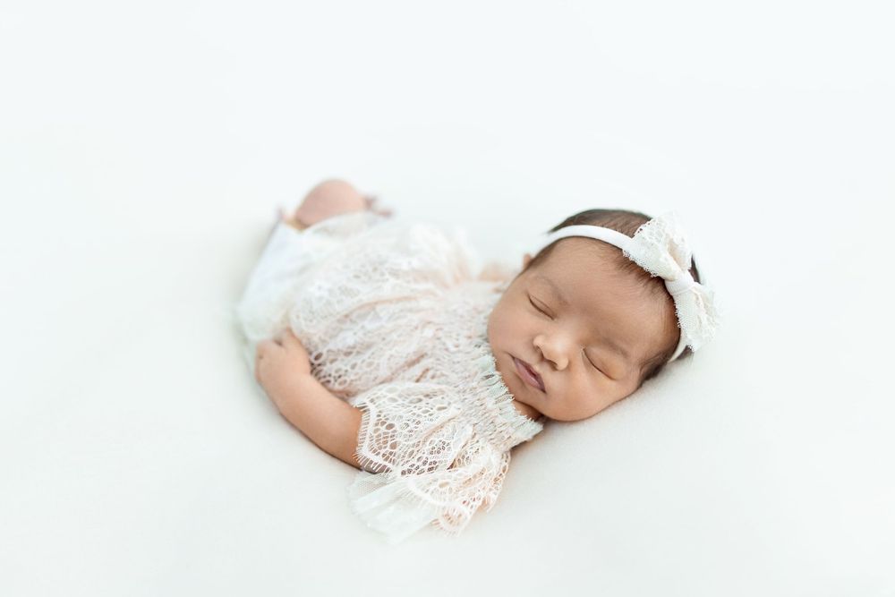A sleeping newborn baby wearing white lace clothing lies peacefully on a soft white blanket.