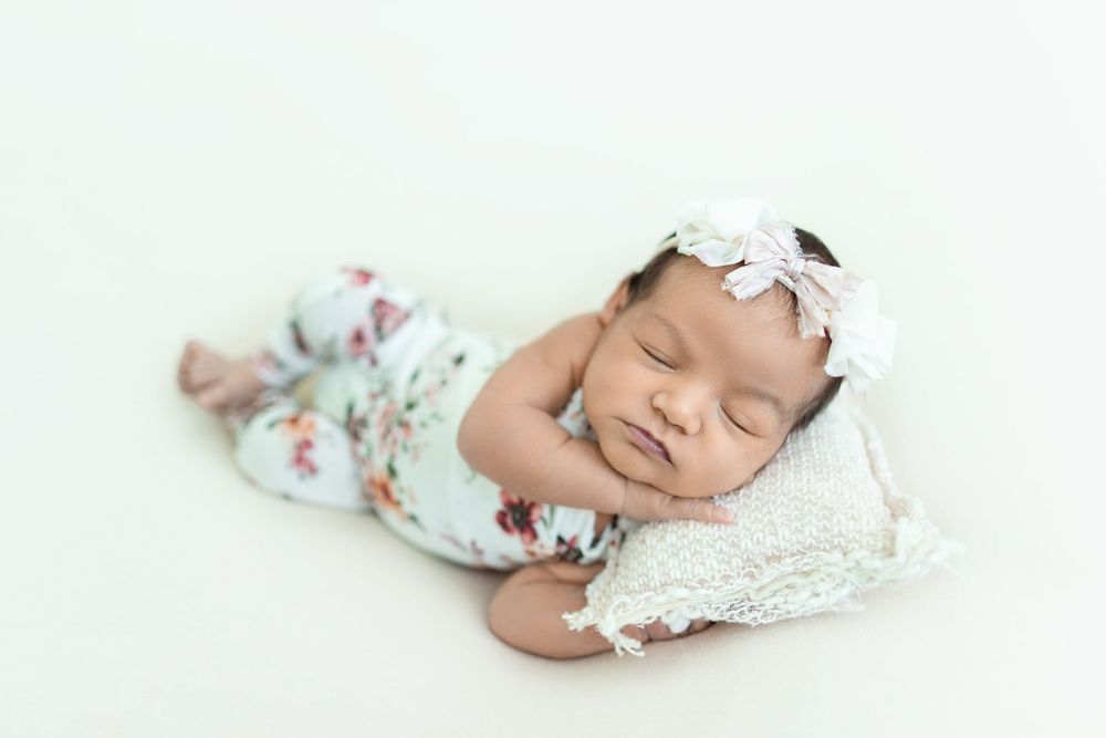 Sleeping newborn baby wearing floral outfit and bow headband lies peacefully on white blanket.