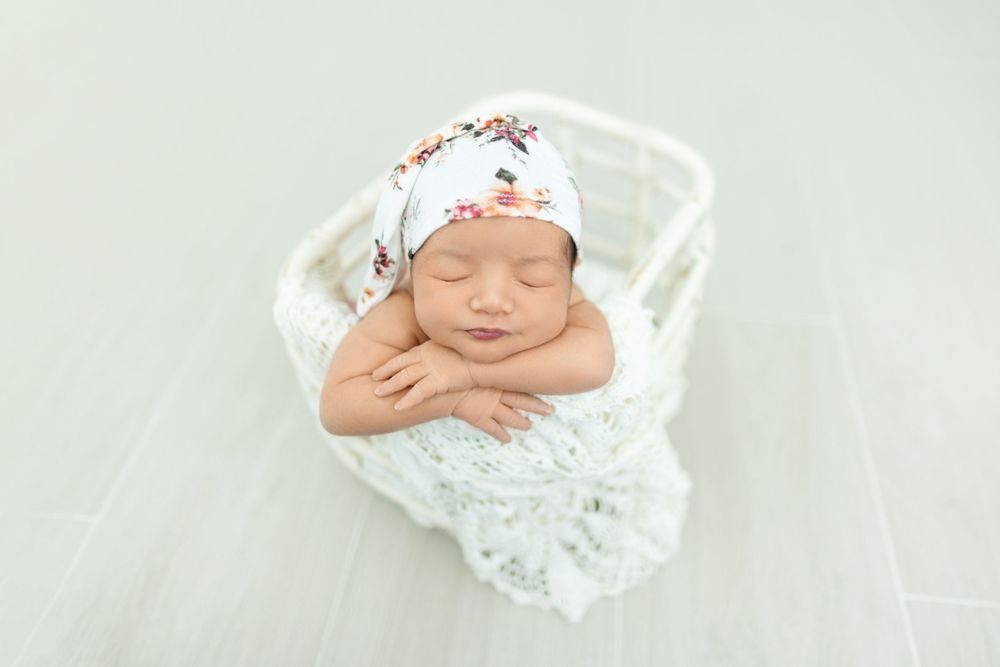 A sleeping newborn swaddled in white lace with a floral headband rests peacefully on a light backdrop.