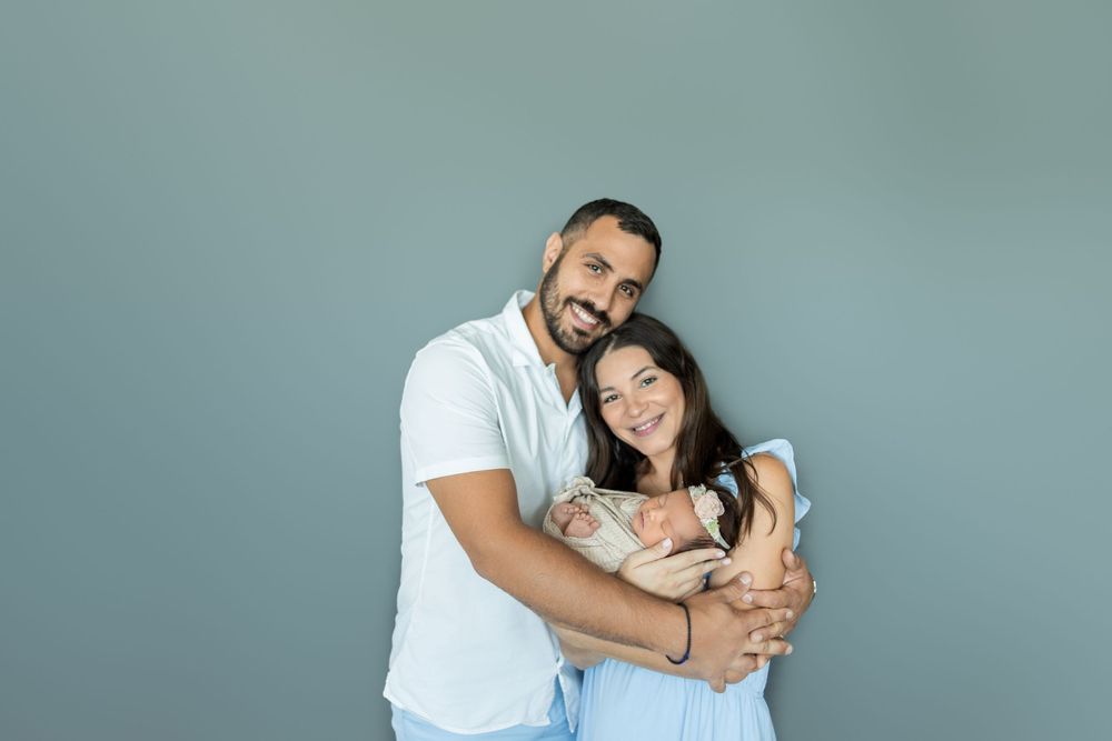 A couple in white outfits embraces while taking selfies together against a teal colored wall.