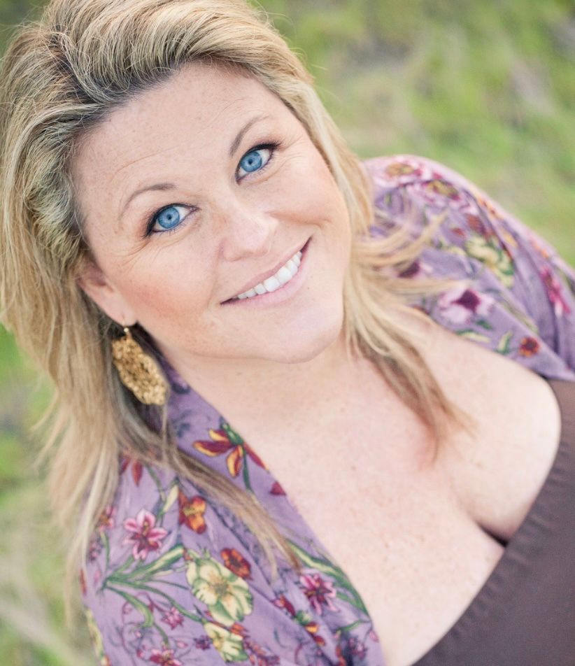 A person in a white top smiles and looks down while posing against a blurred outdoor background.