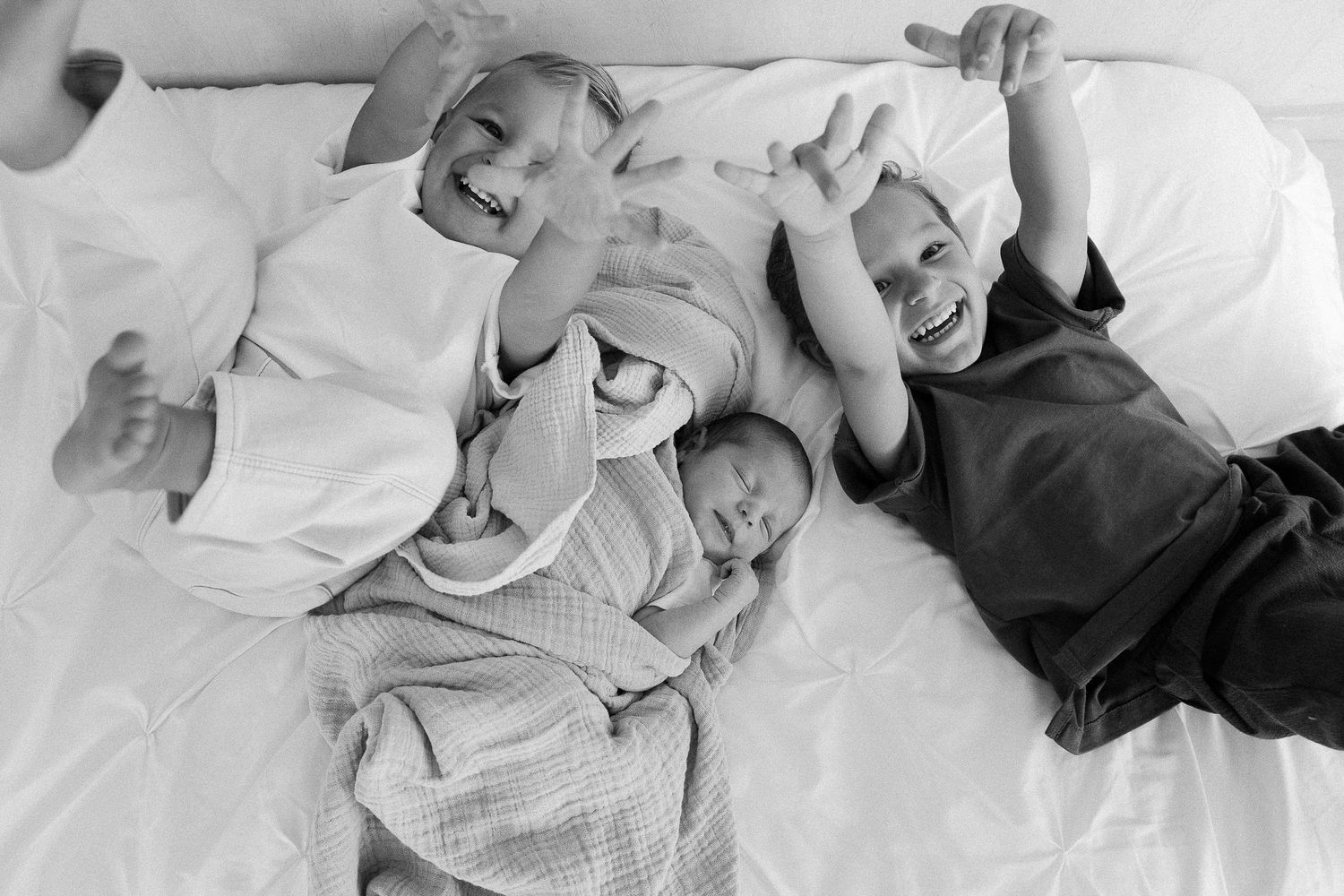 Black and white photo of three siblings playfully laying together on white bedding.