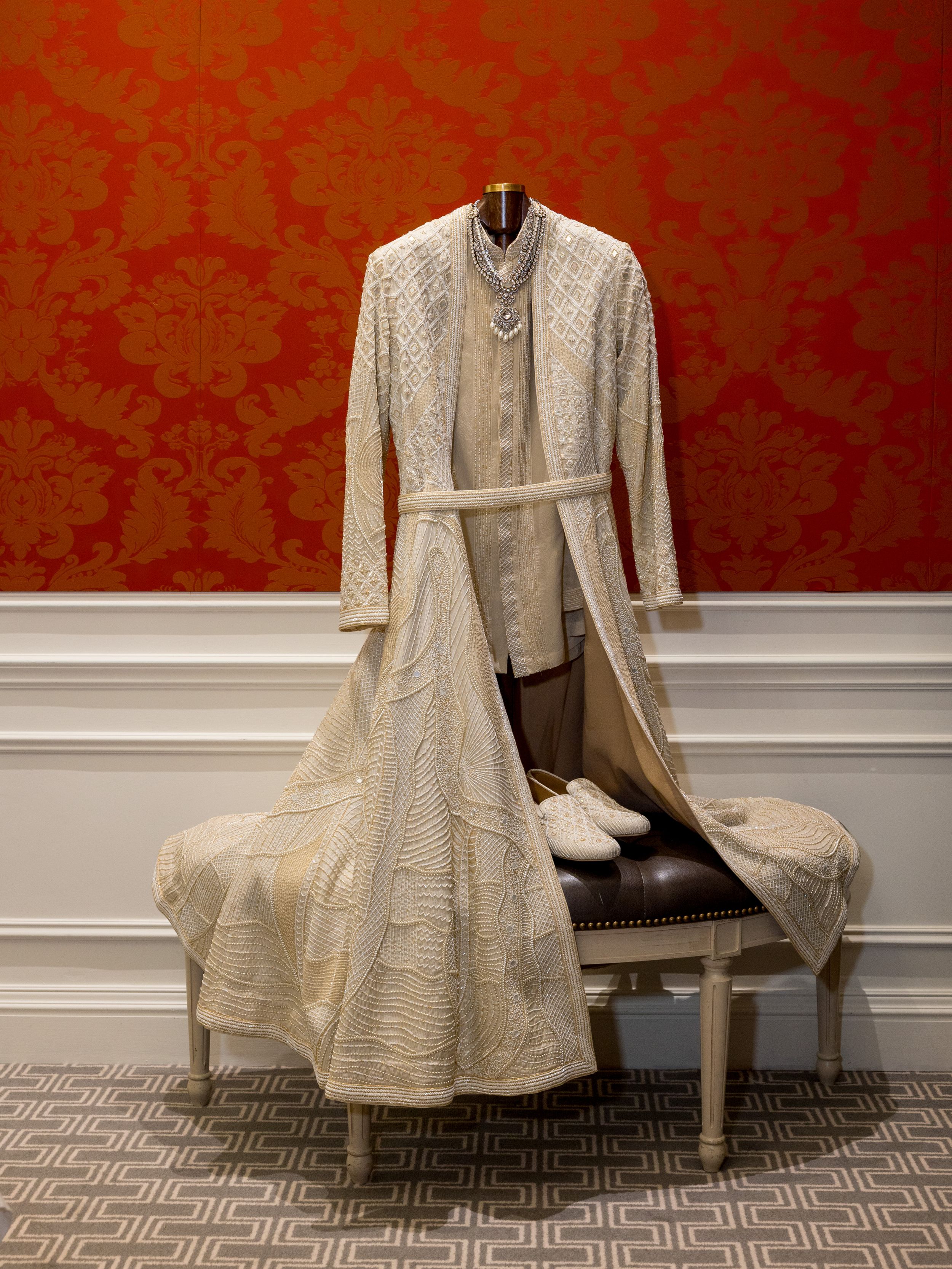 A vintage-style cream colored formal dress displayed against a red damask wallpaper.