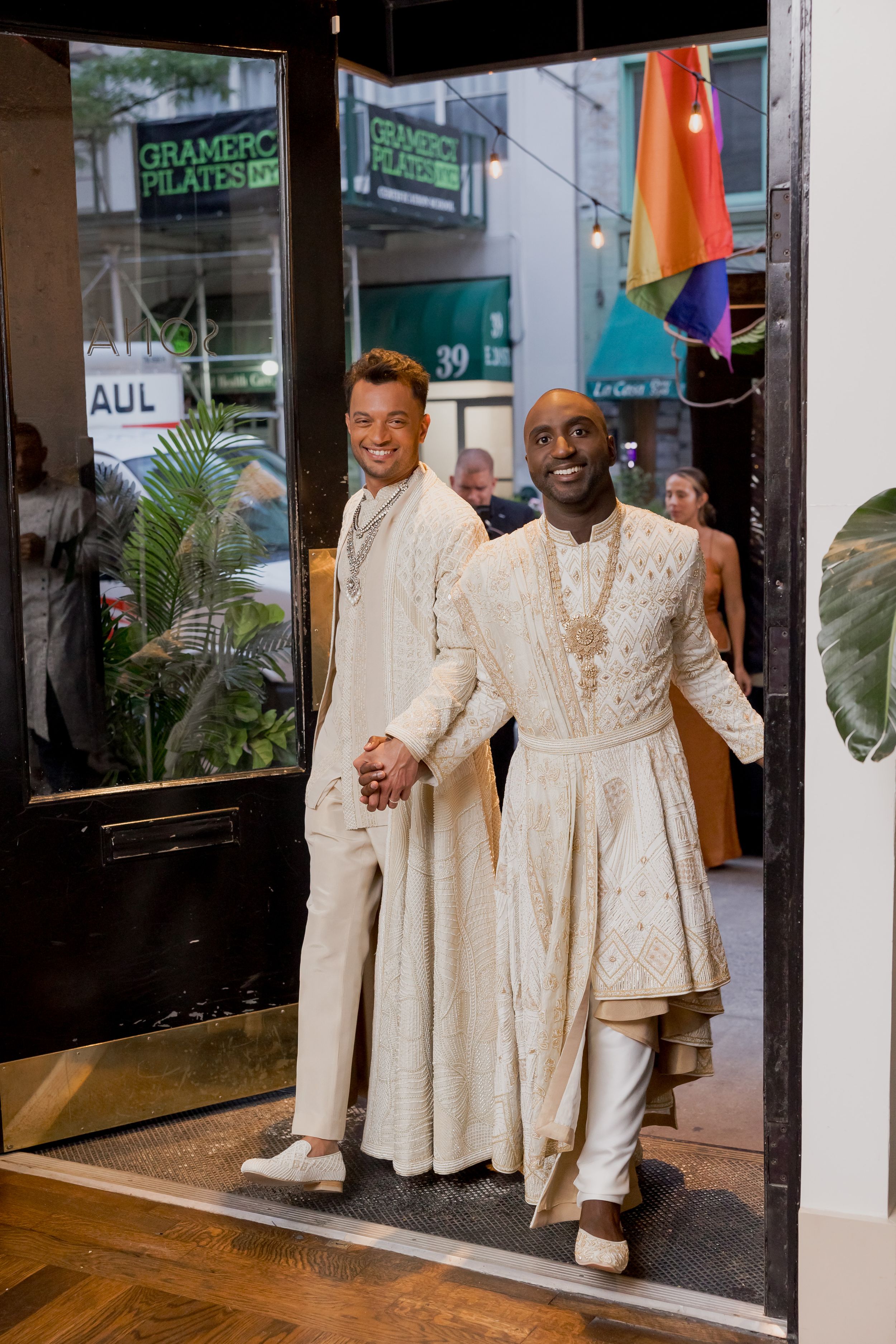 Two people in matching white formal attire standing in a doorway with a pride flag visible in the background.