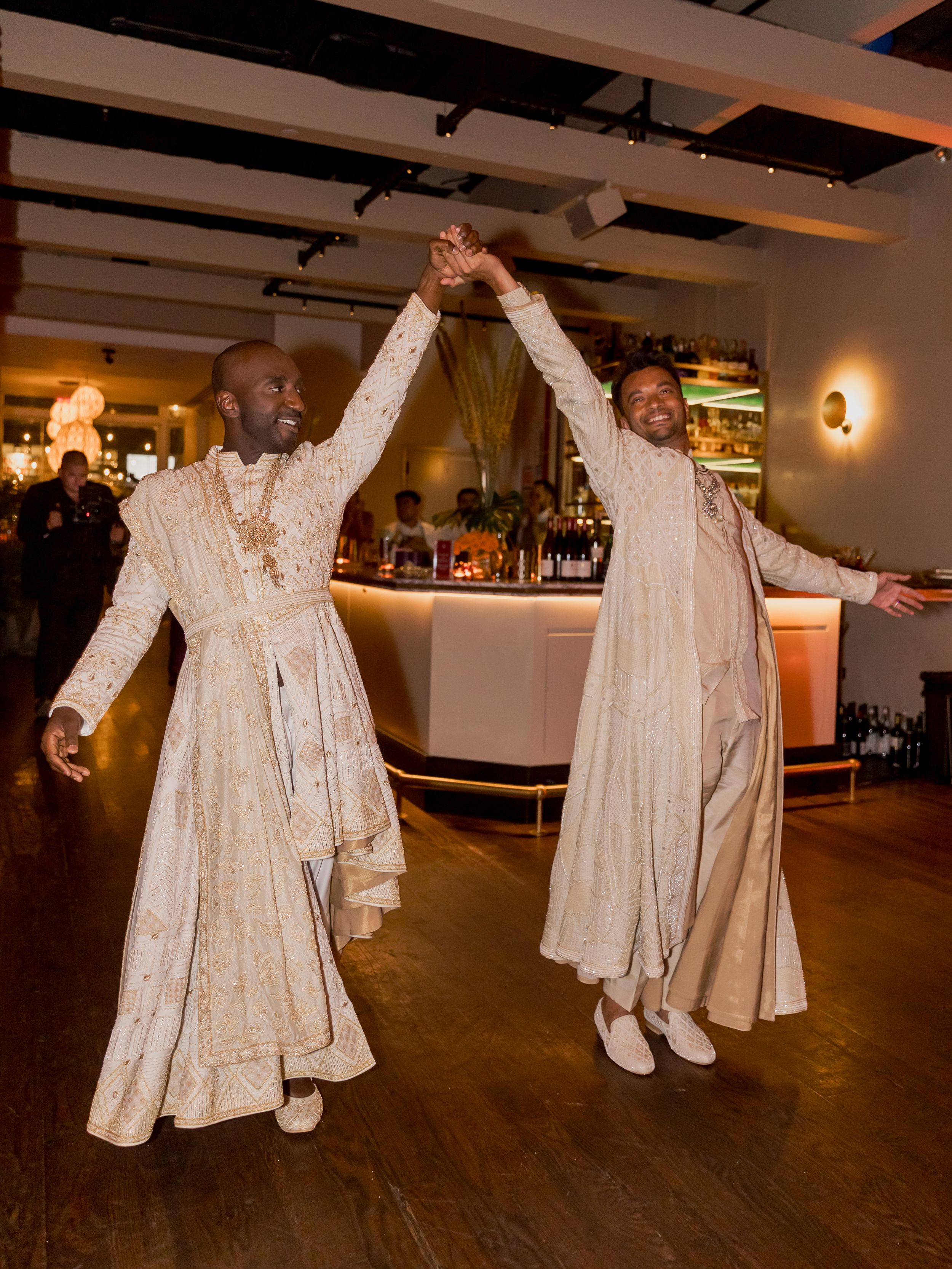Two people in matching white formal attire dancing and holding hands in celebration.