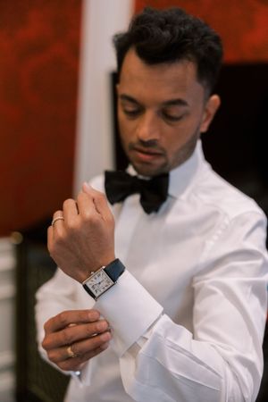 A close-up shot of hands adjusting a watch and cufflink on a crisp white tuxedo shirt.