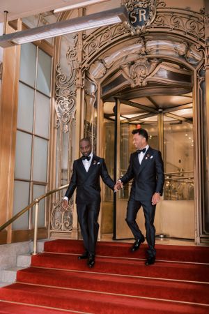 Two people in formal black tuxedos walk through an elegant hallway with ornate gold decor and red carpeting.