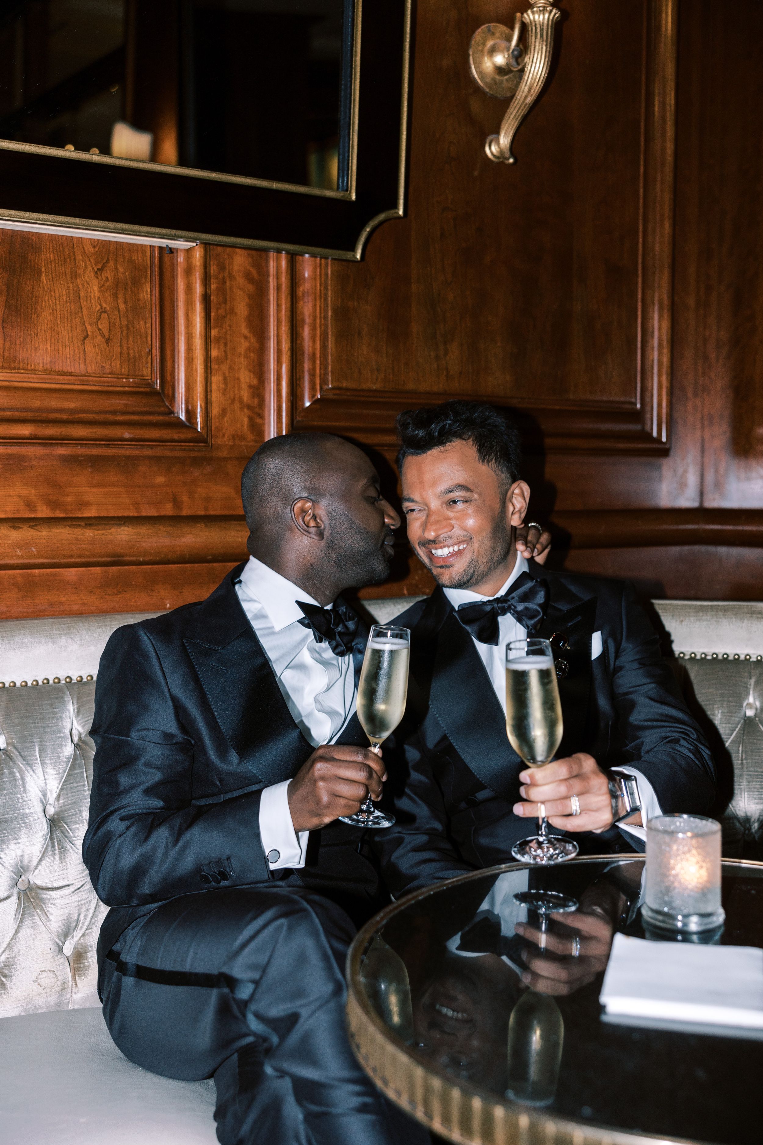 Two people in tuxedos share champagne and intimate moments in an upscale lounge setting.