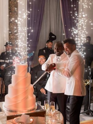 Wedding couple cuts their elegant white tiered cake while guests look on with sparklers in background.