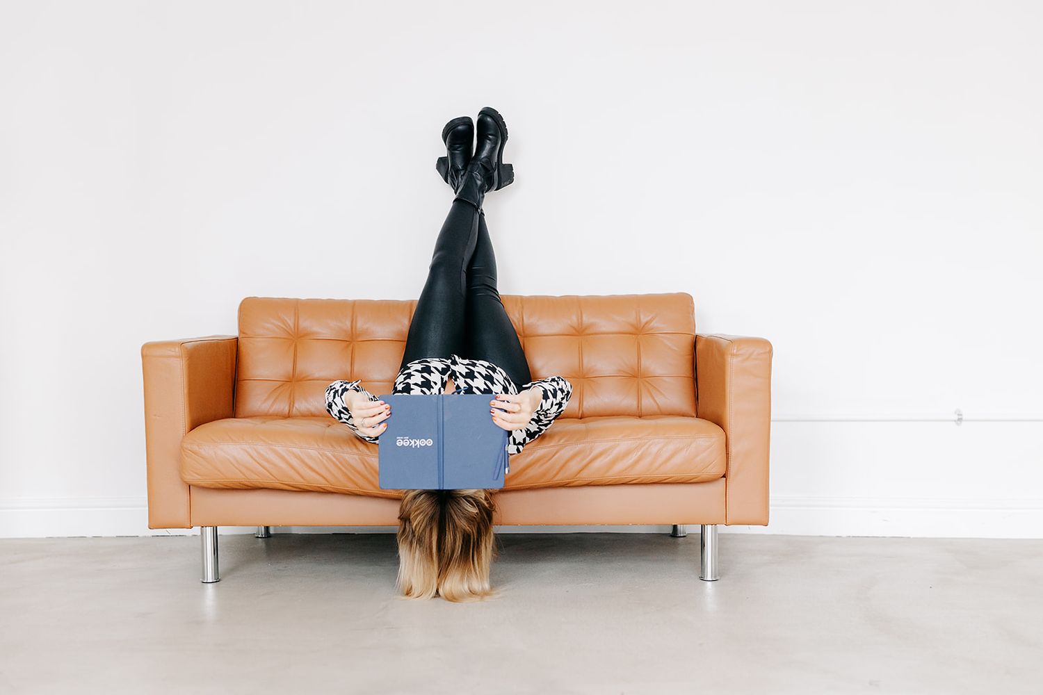 A person wearing a black dress lies face down on a tan leather couch against a white wall.