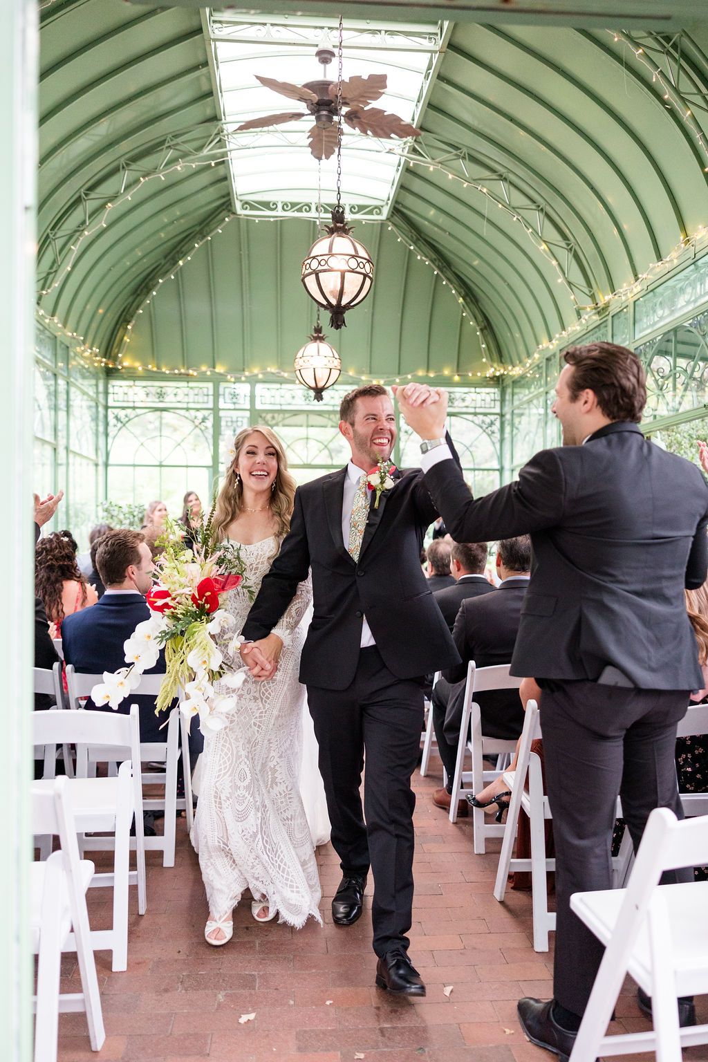 A wedding reception takes place in an elegant conservatory with arched glass ceilings and white folding chairs.