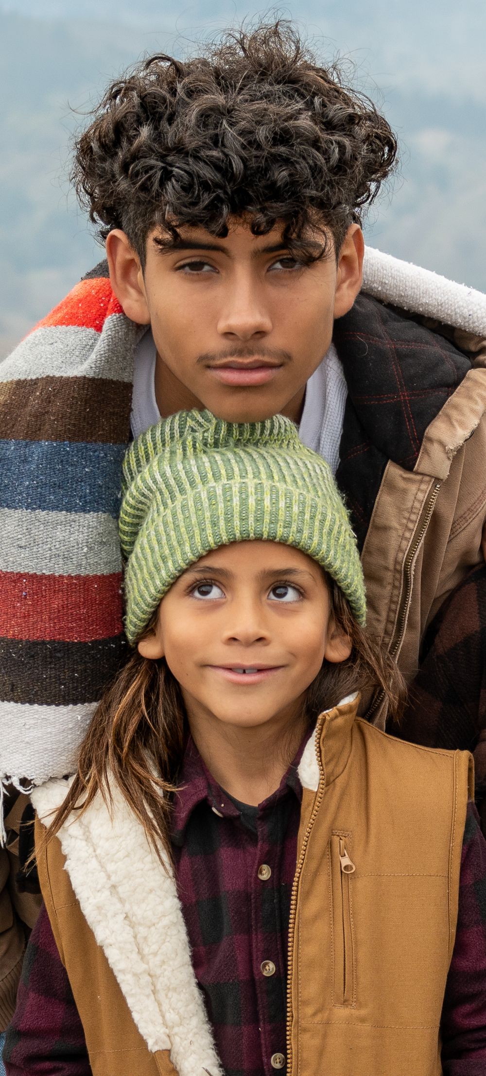 Two people wearing winter clothing including a green knit beanie and brown jacket pose together in a casual portrait series.