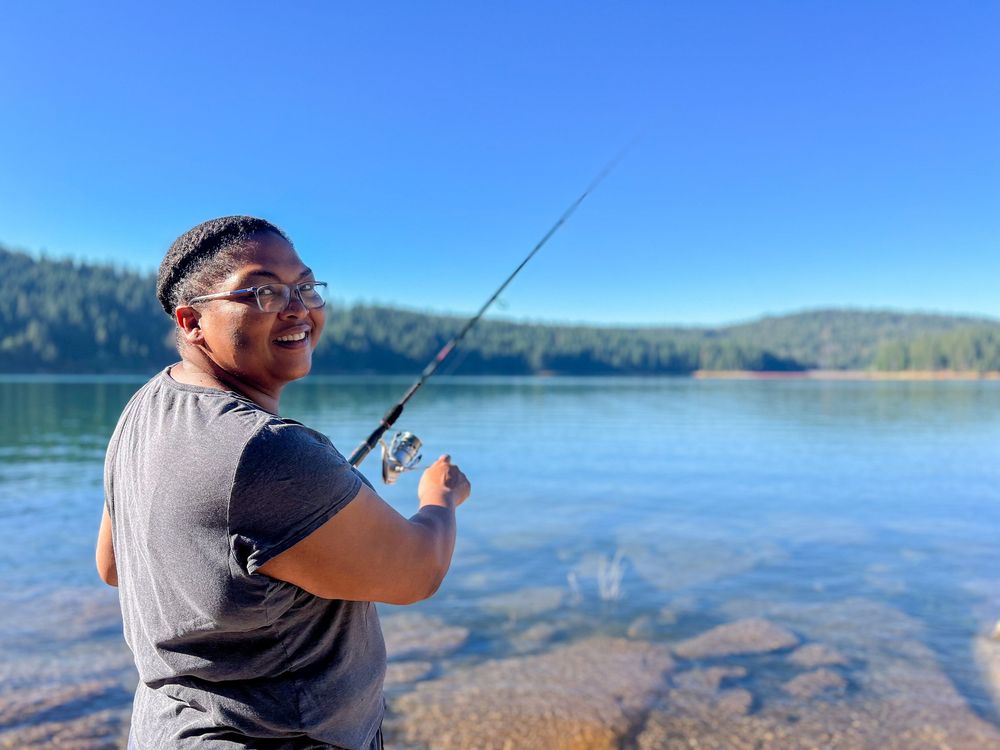 People fishing on a calm lake with mountains and forest in the background.