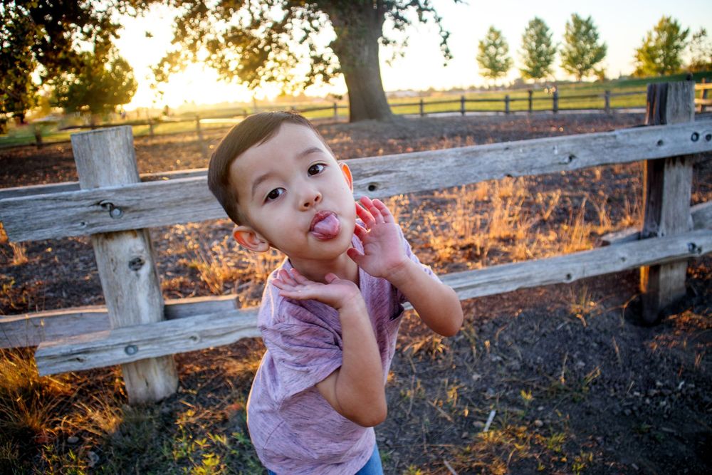 Family photoshoot with child making playful faces near a wooden fence at sunset.