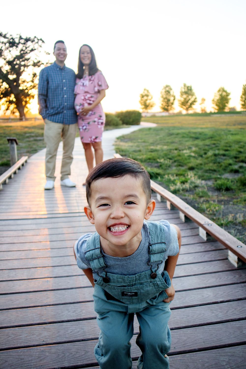 Playful child in denim overalls making funny faces on a wooden boardwalk at sunset.