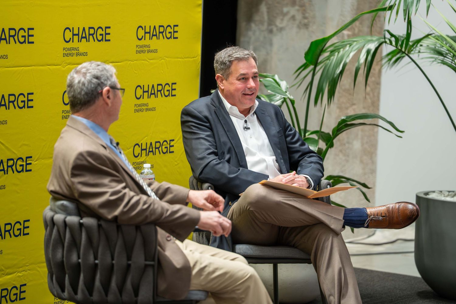 Two people sit in leather chairs having a conversation at an event with yellow CHARGE branded backdrop and potted plants.