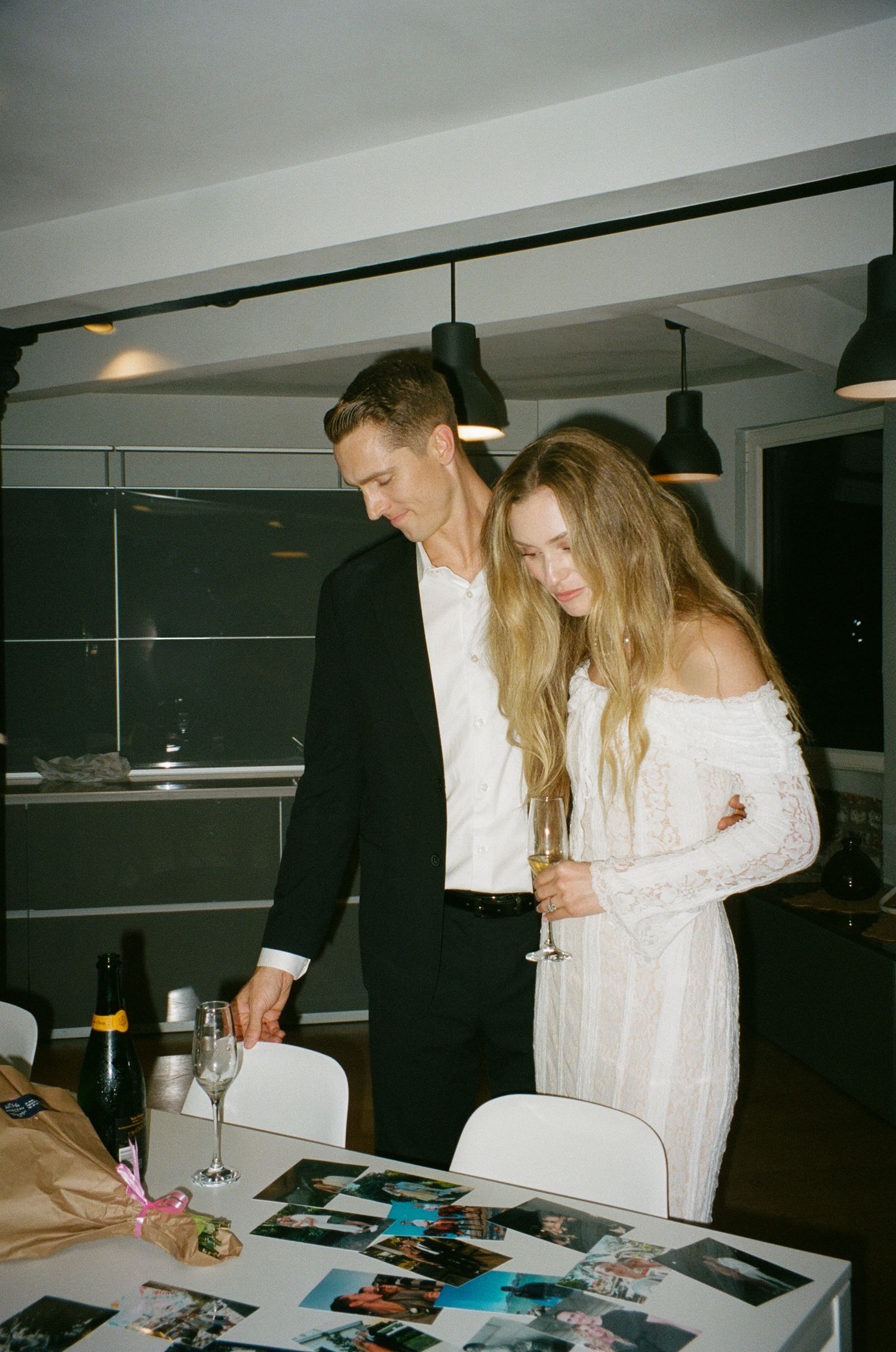 Two people in white clothing examine photographs and documents spread across a table in a dimly lit room.