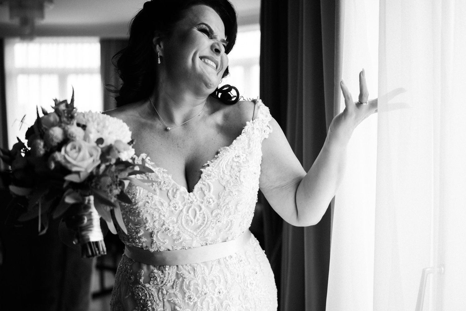 A bride in a white lace wedding dress laughs joyfully during a candid moment captured in black and white photography.