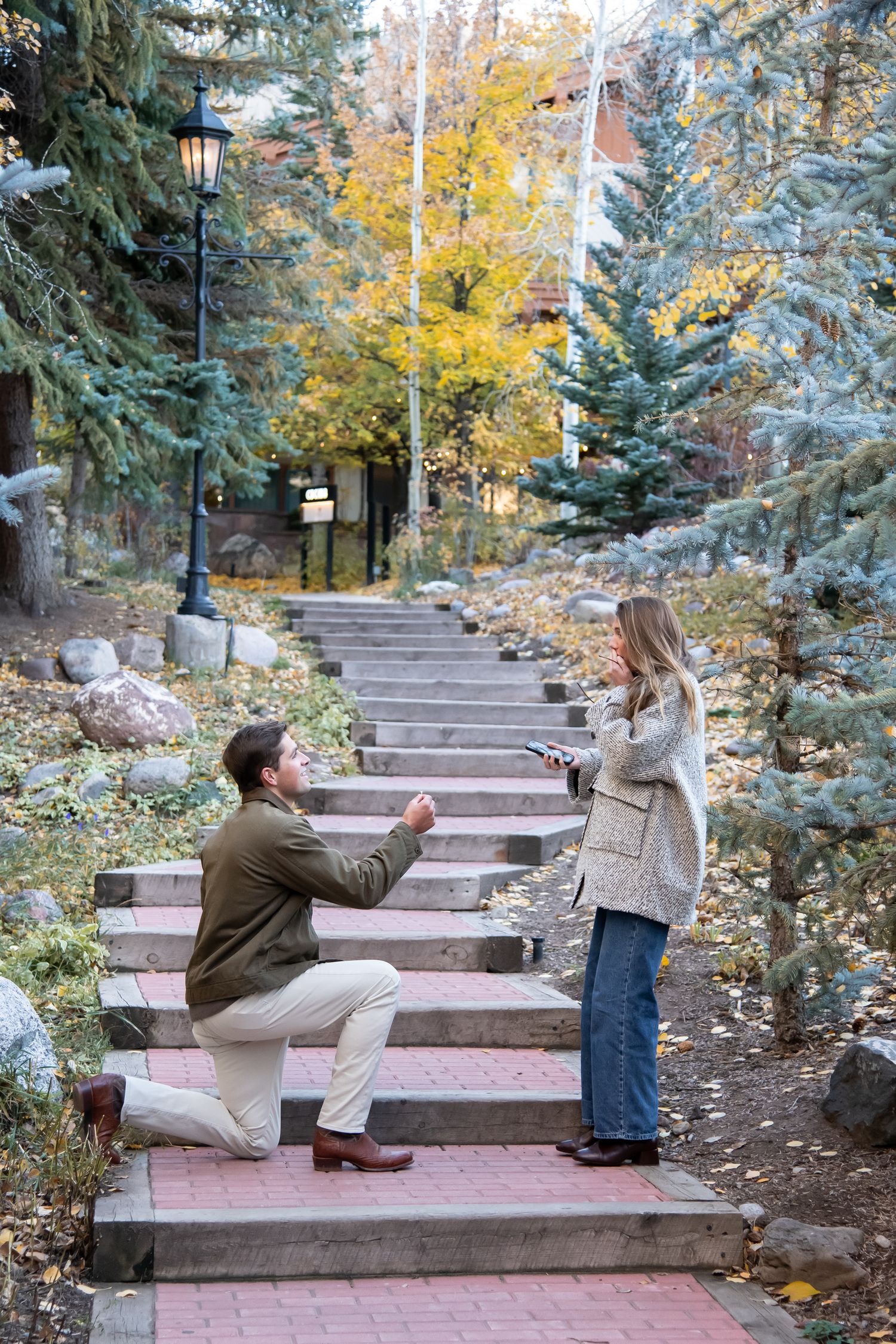 A romantic proposal scene unfolds on outdoor stone steps surrounded by evergreen trees and autumn foliage in Vail Village Colorado.