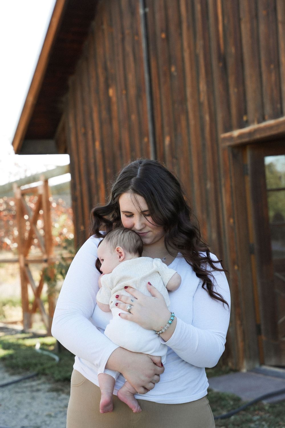 A tender moment between parent and infant is captured in front of a rustic wooden barn on a sunny day.