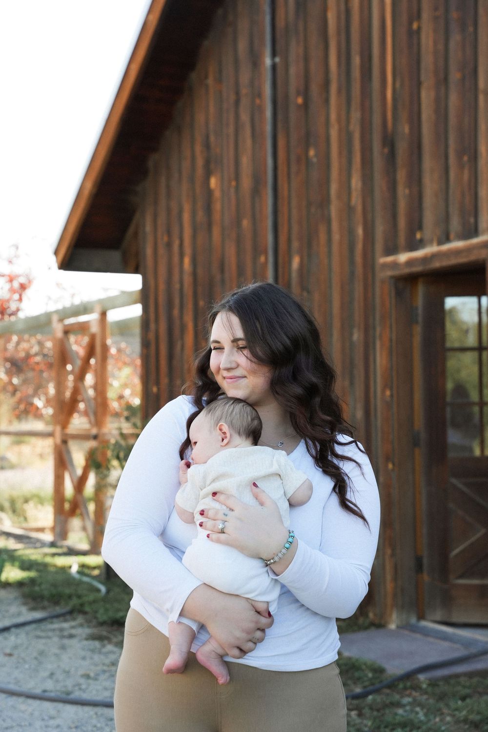 Mother in white sweater holds and plays with baby outdoors near rustic wooden building.