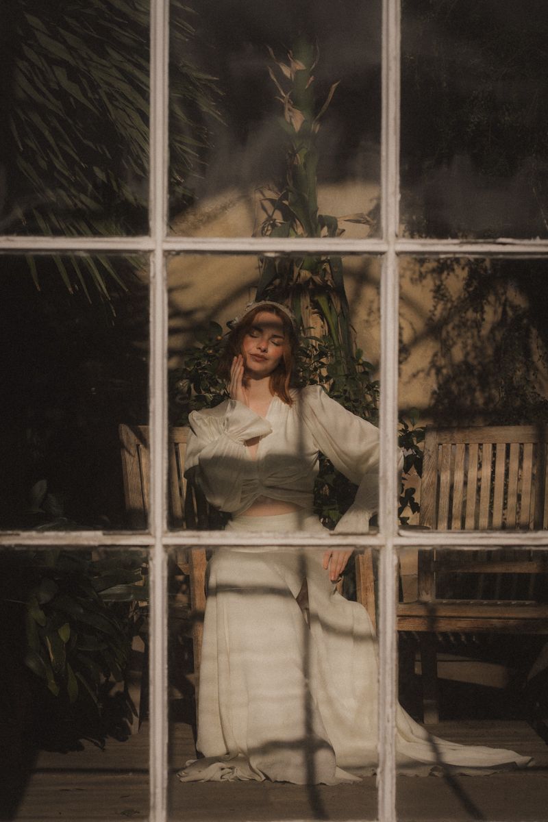 A person in a white sweater poses by a window with dramatic shadows creating an artistic portrait effect.