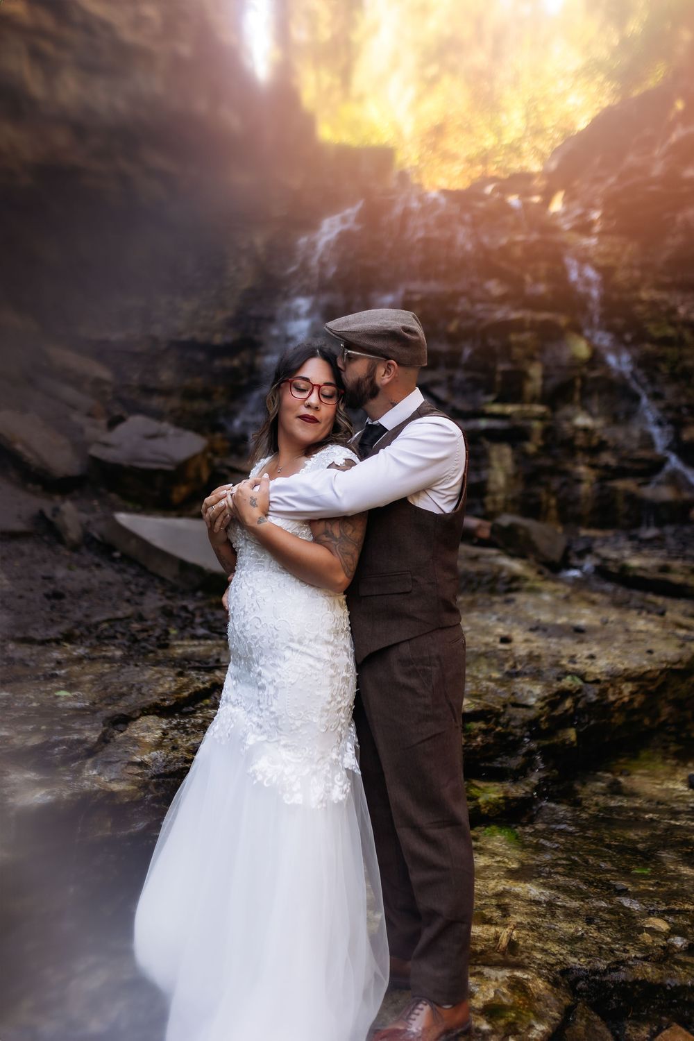 A romantic wedding photo of a couple embracing in front of a misty waterfall at sunset.
