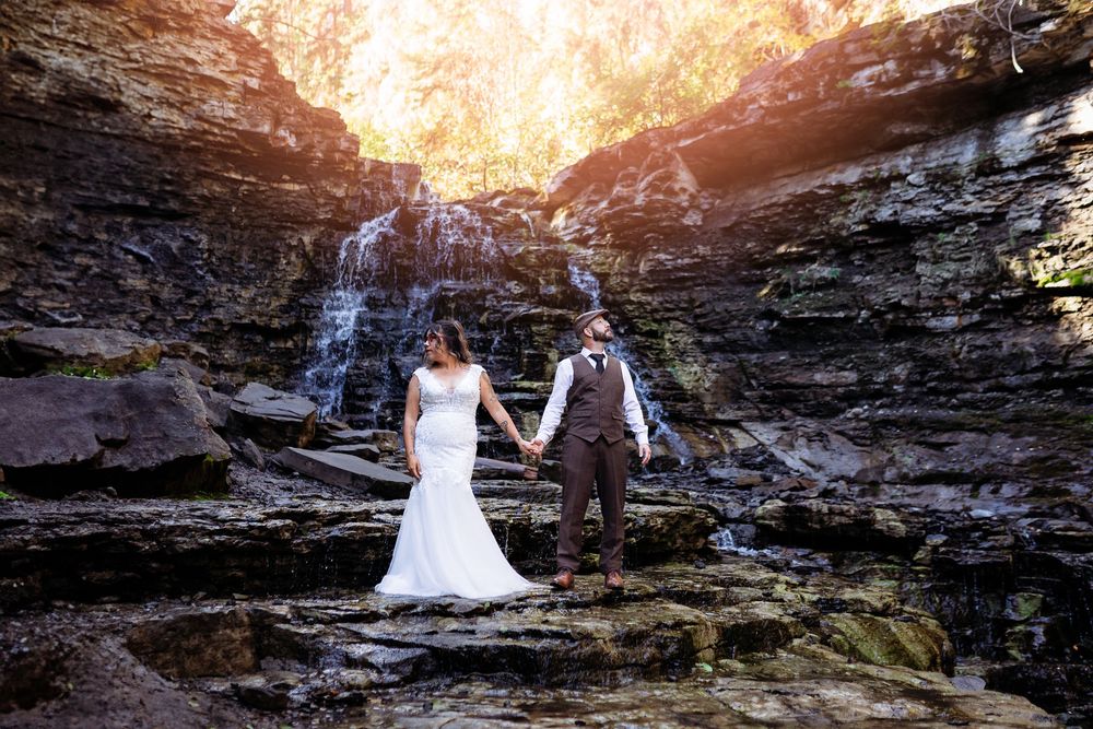 A couple in wedding attire poses against dramatic rocky cliffs during golden hour sunset.