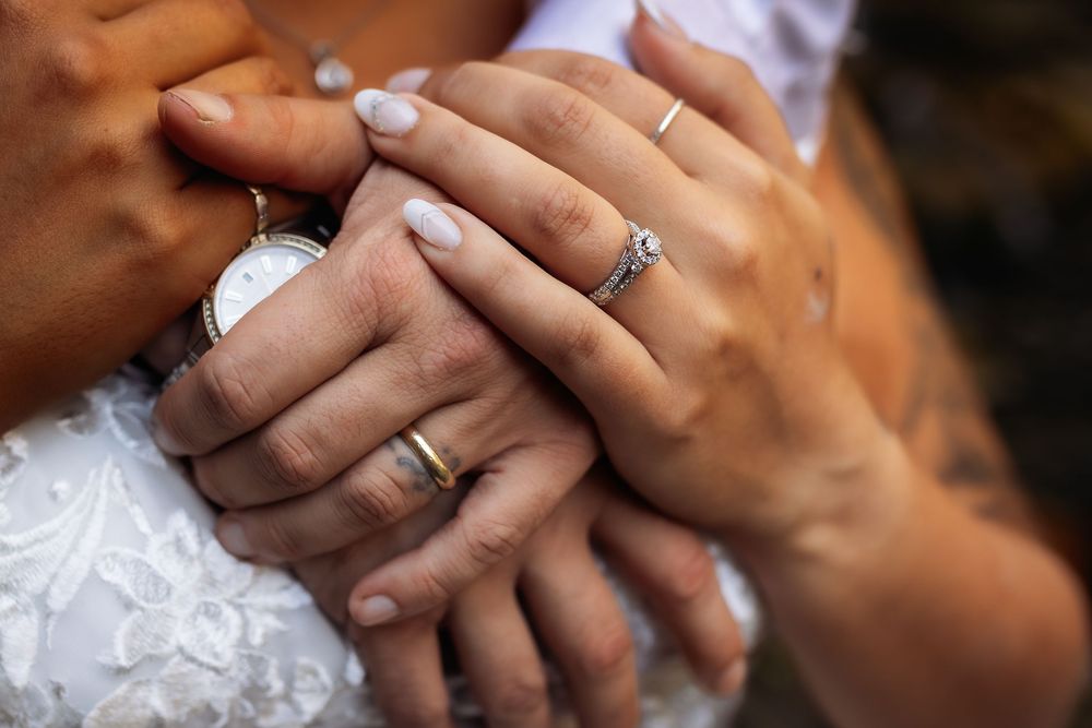 Close-up photo of hands with wedding rings showing bride and groom's fingers intertwined against white lace.