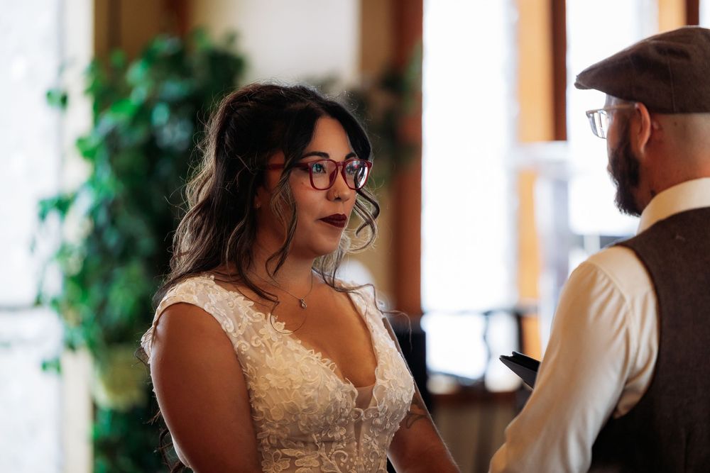 A bride in glasses and a lace wedding dress stands indoors during a wedding ceremony.