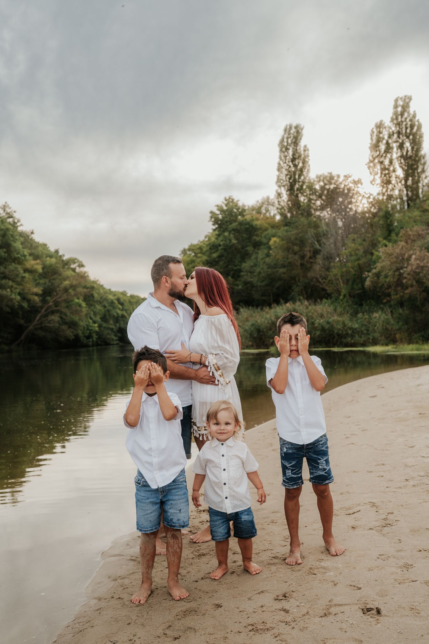 Family wearing white and denim stands together on sandy beach near lake under cloudy sky.