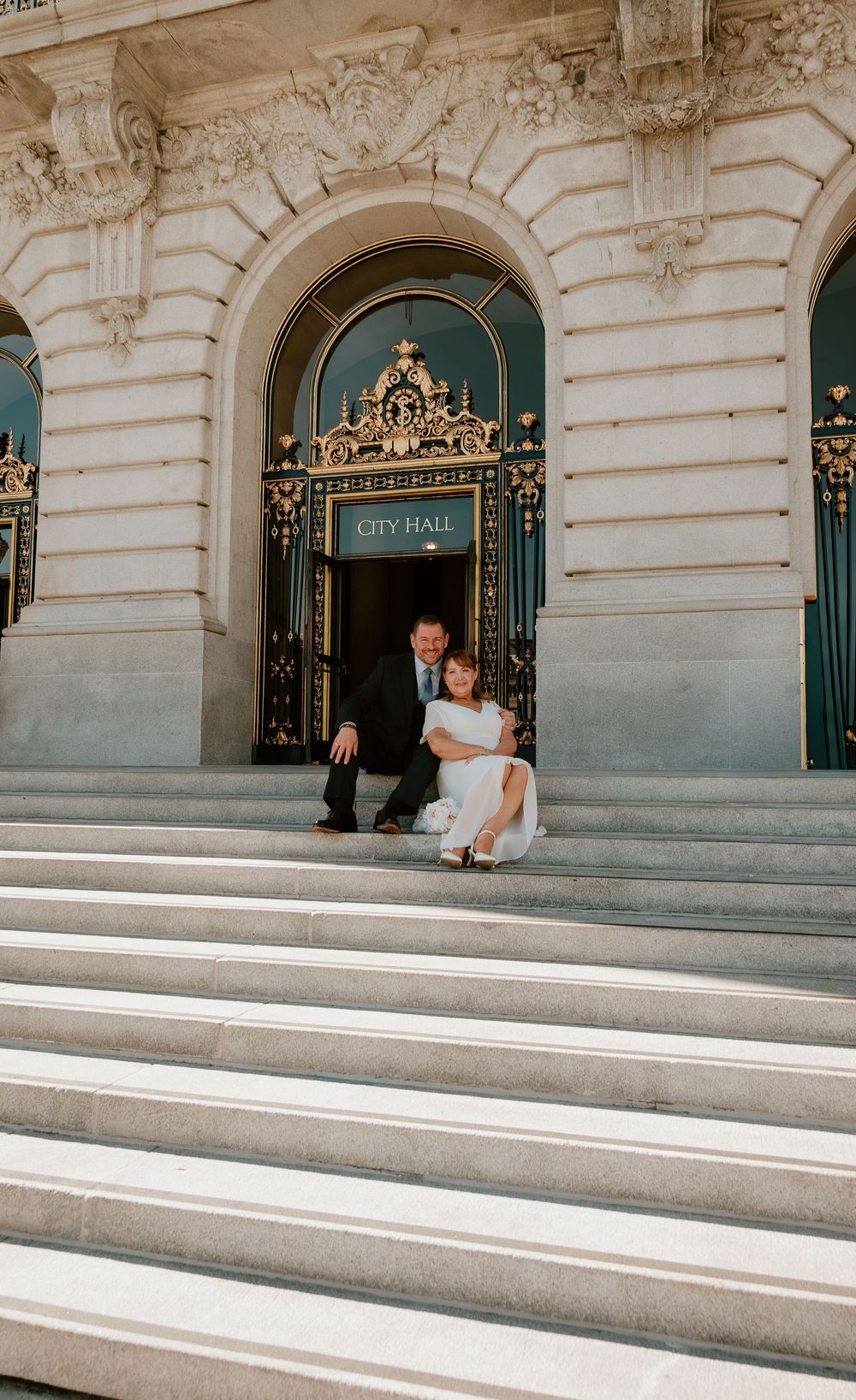 A couple sits on the grand stone steps of a historic city hall building with ornate architectural details.