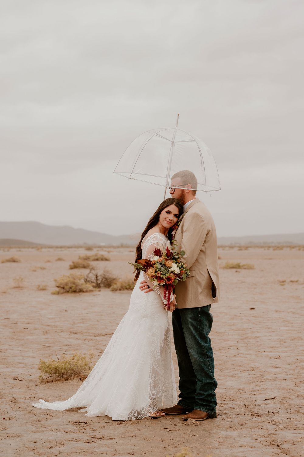 A couple in wedding attire shares an intimate moment on a desert landscape under an overcast sky.