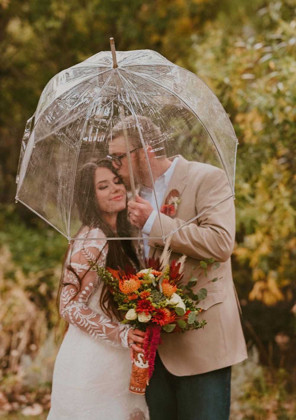 A couple shares an intimate moment under a clear umbrella while holding vibrant orange and red flowers in an autumn setting.