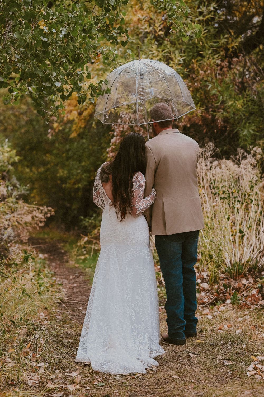 A couple in wedding attire walks through a wooded path during autumn with golden leaves surrounding them.