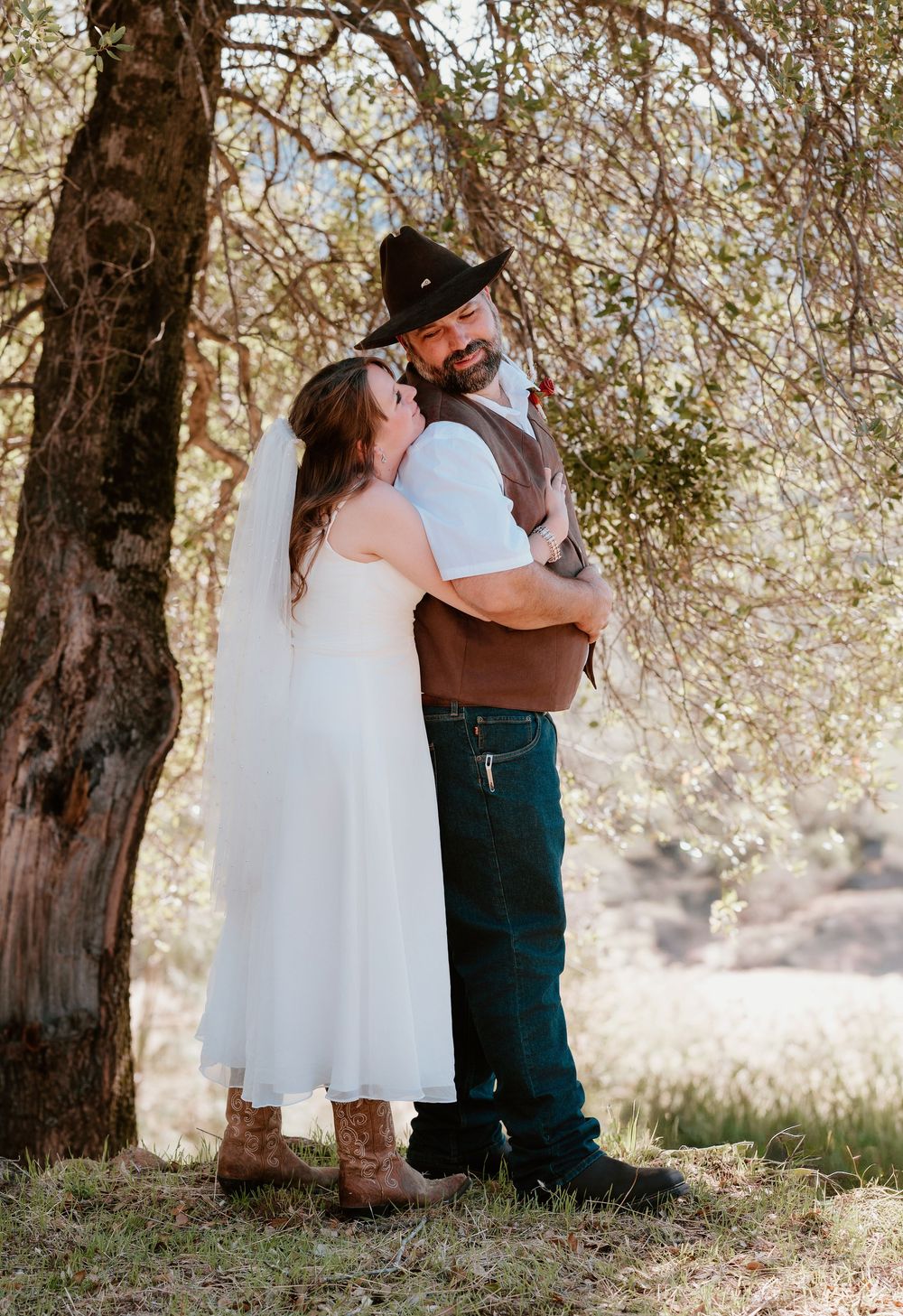 A romantic outdoor wedding moment under a tree with the bride in white dress and groom in western attire and cowboy hat.