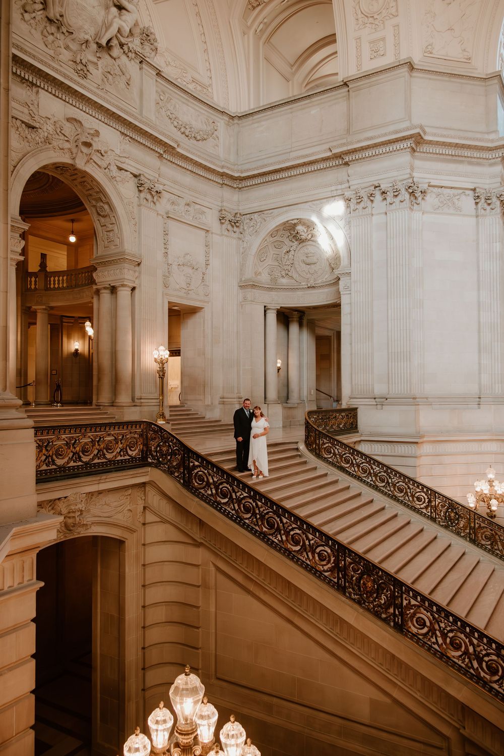 Elegant marble staircase with ornate iron railings and chandeliers in a grand historic building interior.