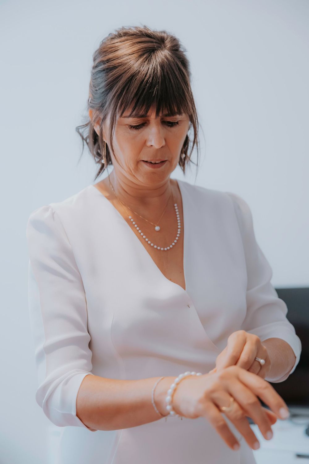 Woman in white blouse adjusts pearl jewelry while sitting against light background.