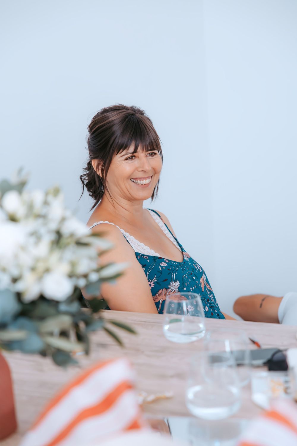 A person in a teal dress sits at a dining table with floral decor and glassware, smiling during a celebration.