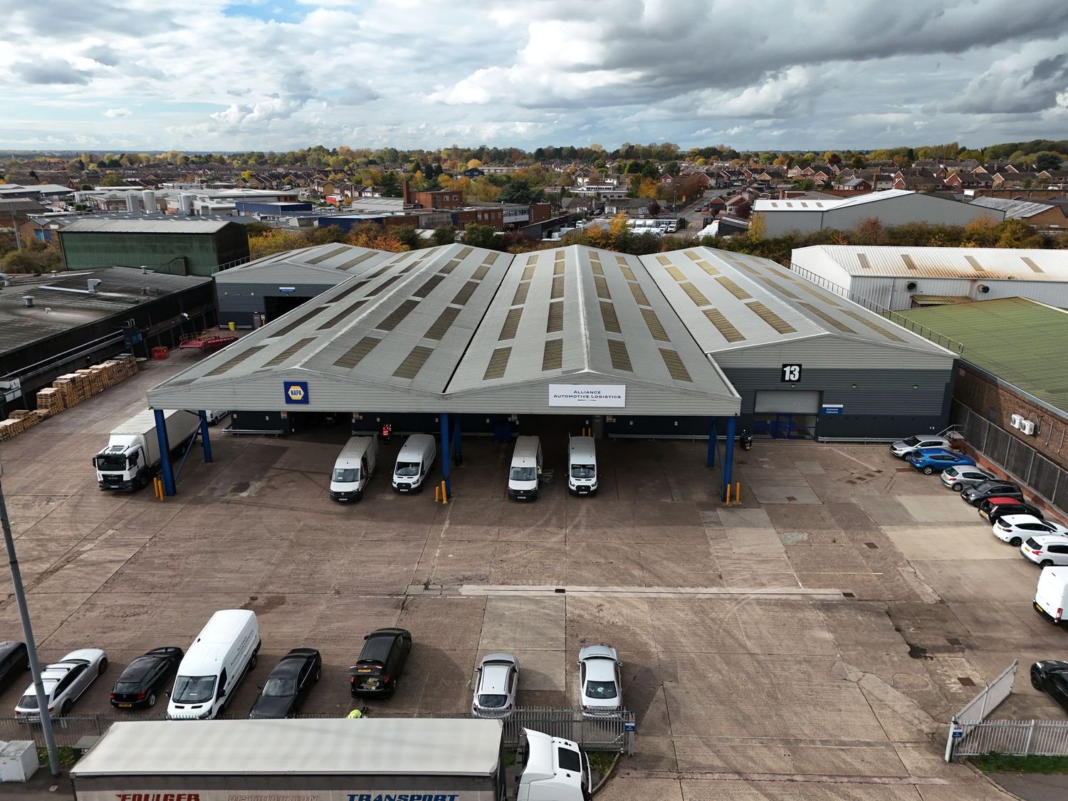 Aerial view of large industrial warehouses and parking lots with trucks and cars under cloudy skies.