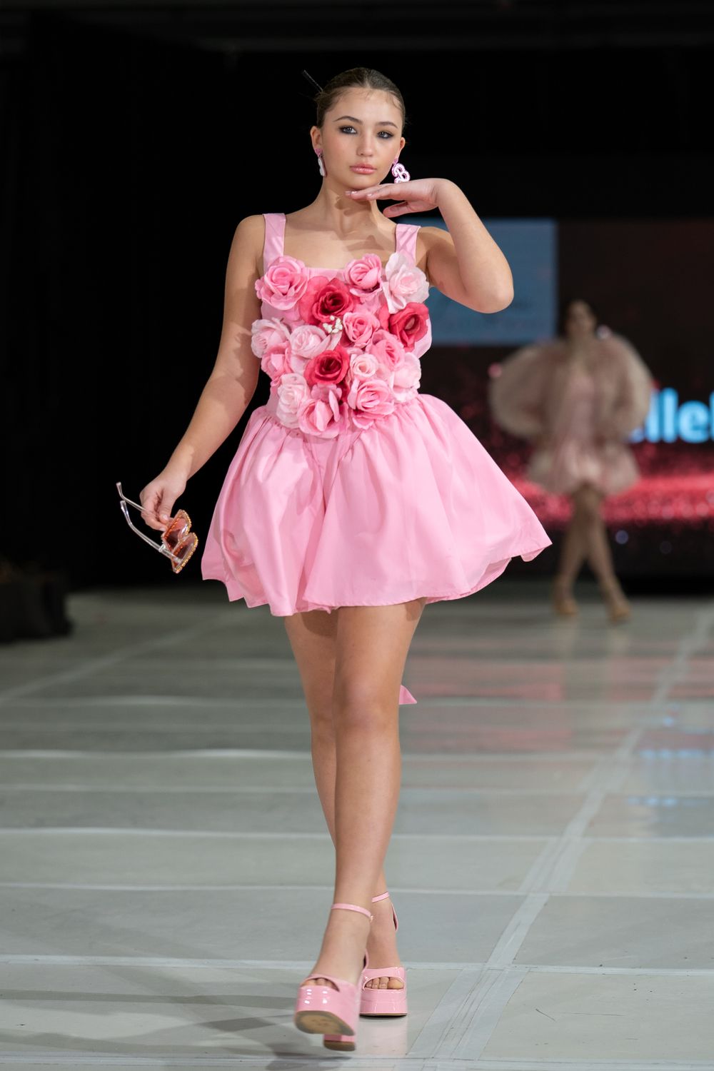 Fashion model walks runway in short pink dress with floral bodice detail and matching platform sandals at fashion show.