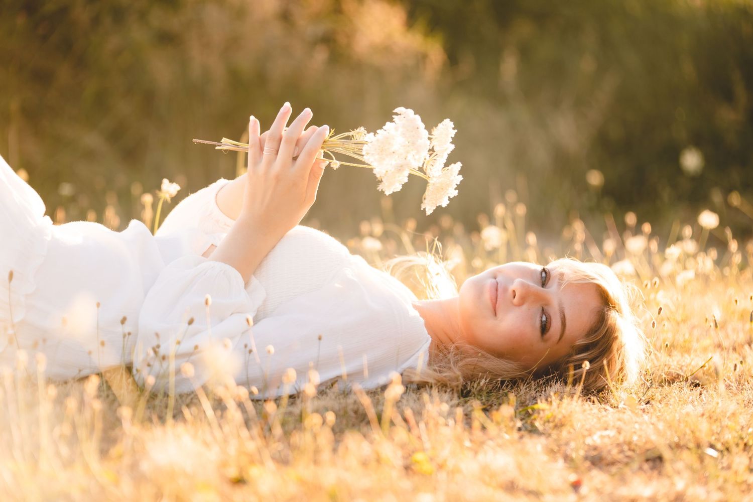 Someone in a white dress lies in sunlit golden grass holding flowers at sunset.