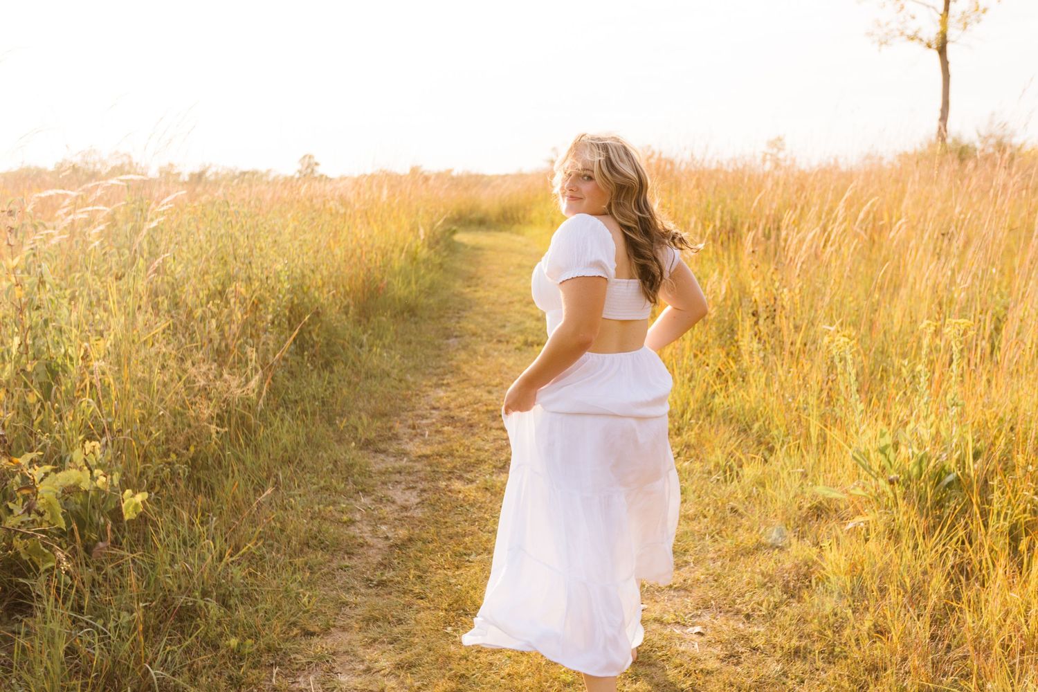 A person in a flowing white dress walks along a sunlit dirt path through tall golden grass at sunset.