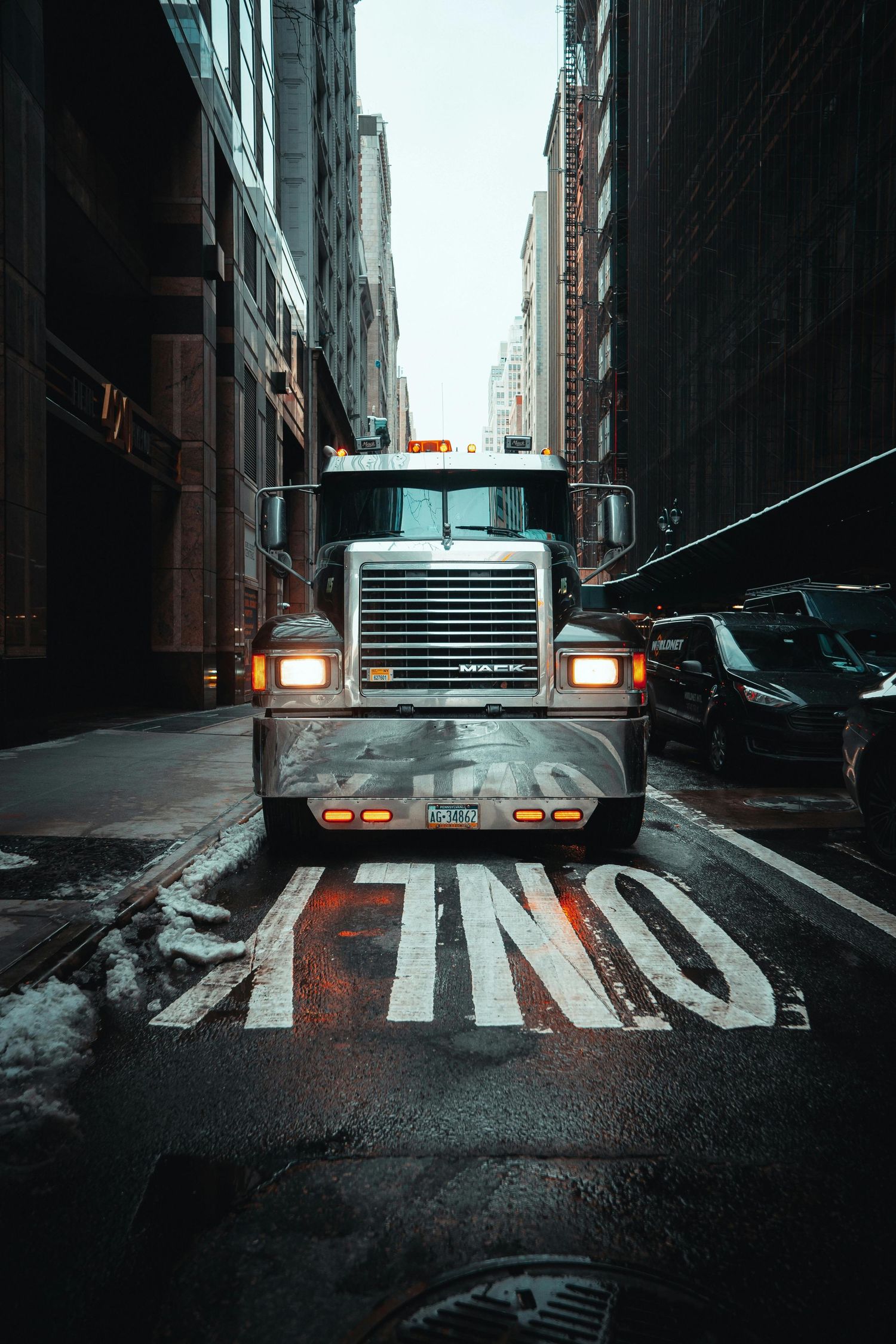 A large semi truck with illuminated headlights parked on a narrow city street with STOP painted on the wet pavement.