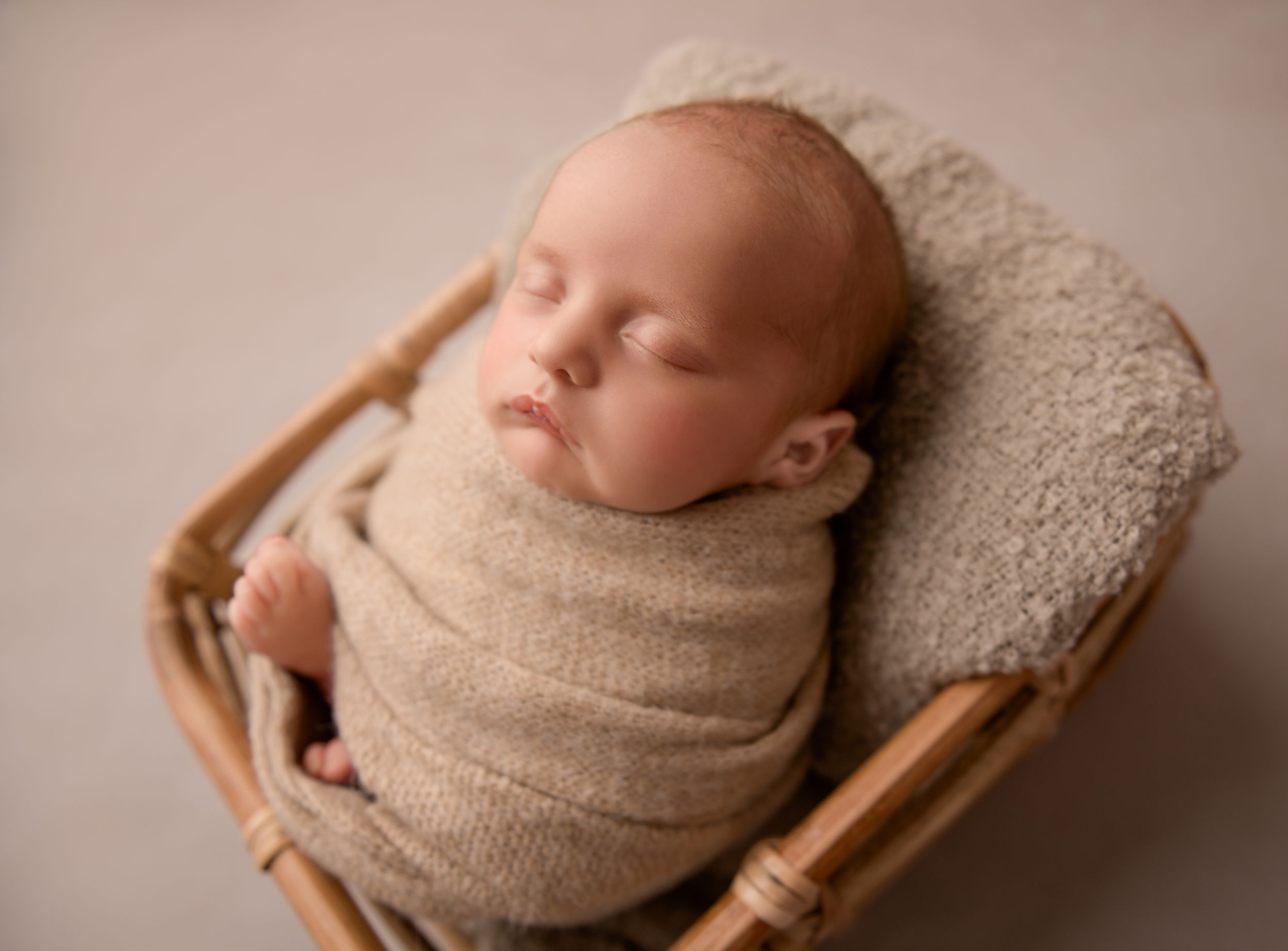 A newborn baby sleeps peacefully whilst being photographed on a wooden floor in a curled up position wrapped in soft, neutral fabric.