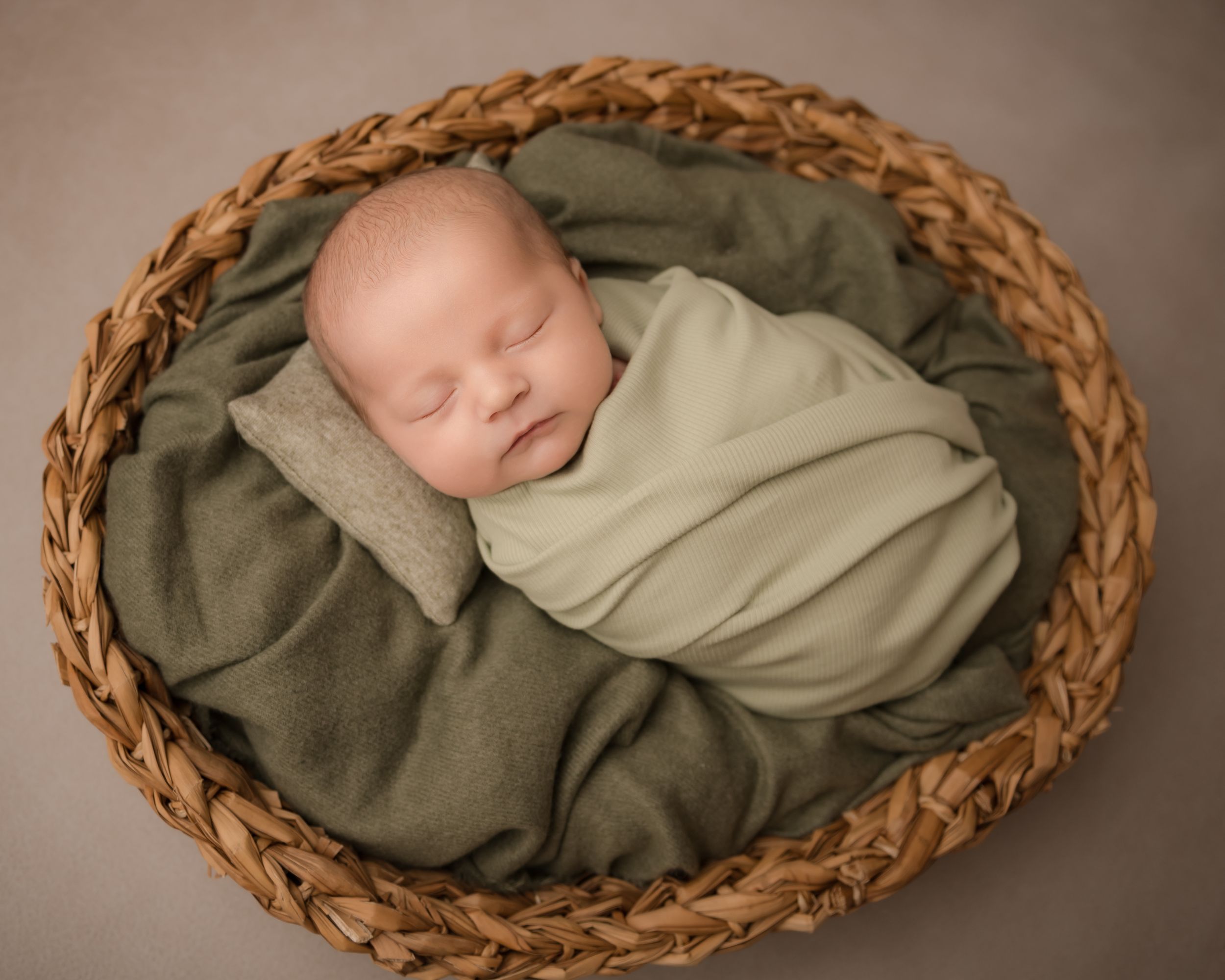 A newborn baby sleeps peacefully whilst being photographed on a wooden floor in a curled up position wrapped in soft, neutral fabric.