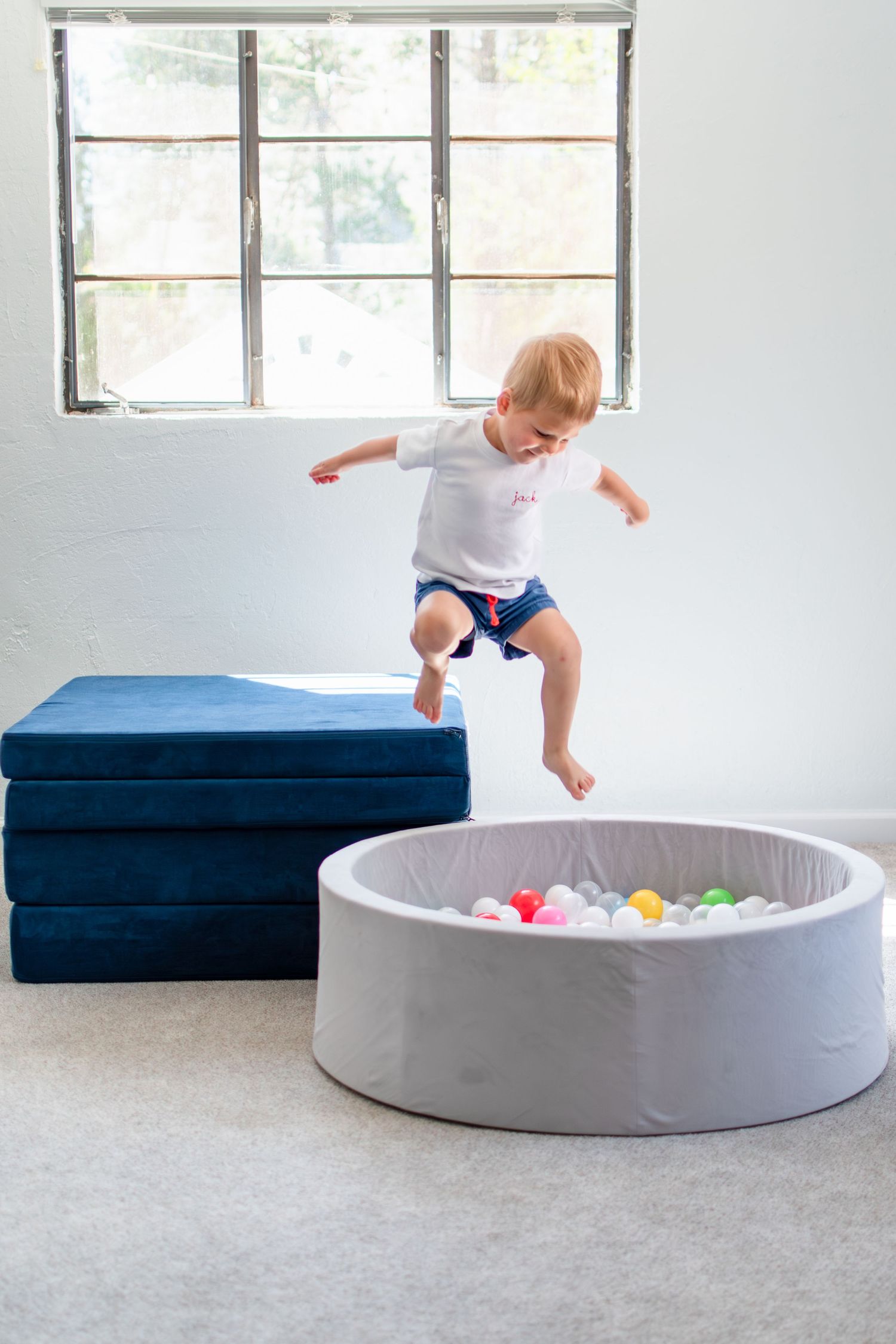 Child in white shirt and blue shorts jumping from blue mats into a gray ball pit in a bright room near a window.