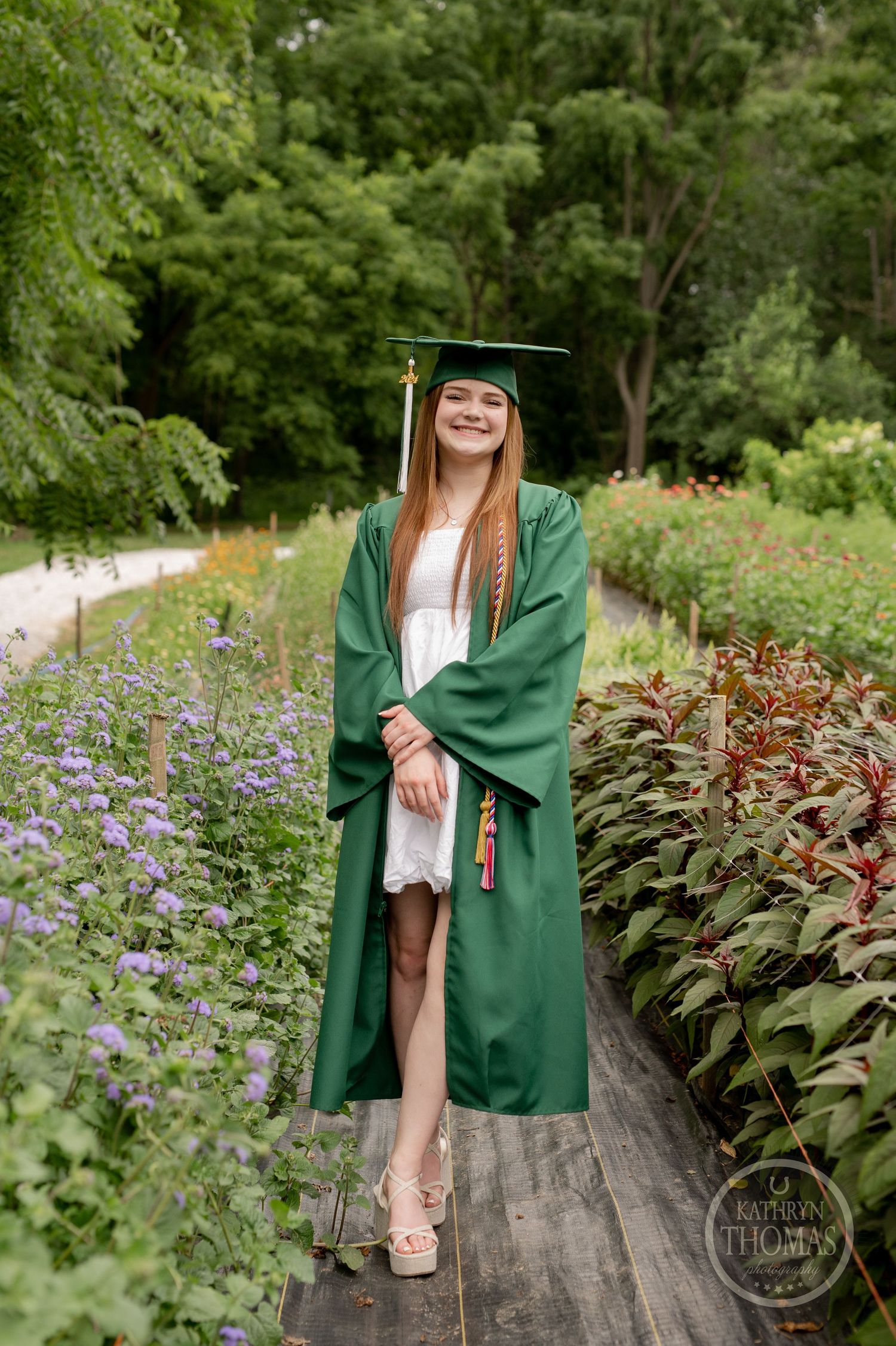 Graduate in green academic regalia stands on wooden pathway surrounded by purple flowers and greenery.
